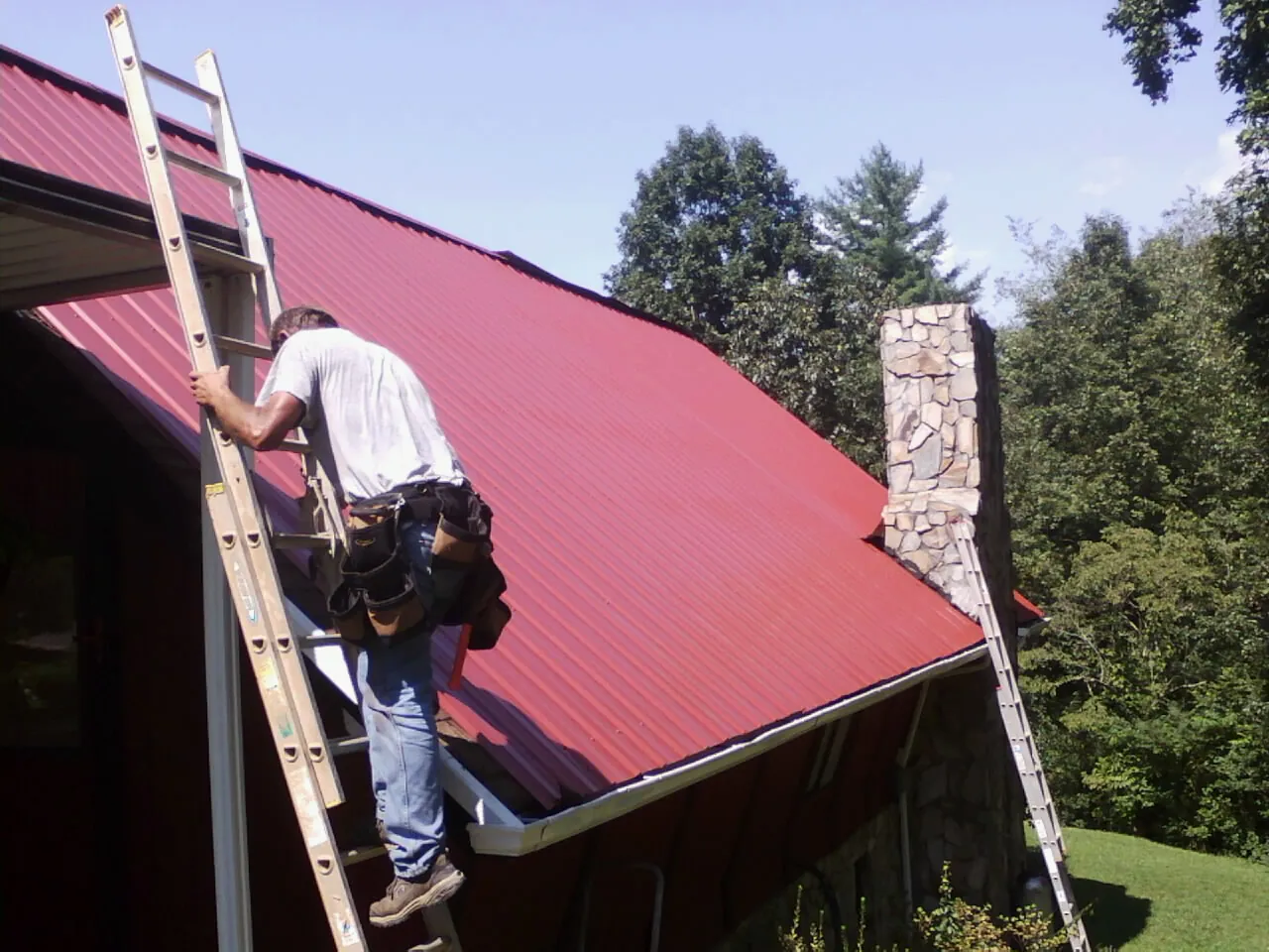 Roofer on a ladder beside a red metal roof and stone chimney — Soffit & Fascia Repair by Roofing Brothers in Stoughton